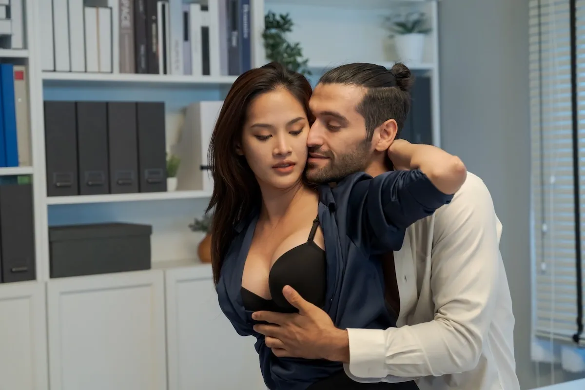 Man intimately embracing a woman in a partially unbuttoned blouse inside an office setting, symbolizing temptation behind closed doors.