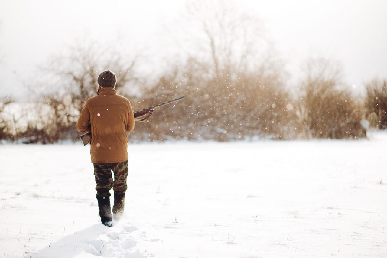 Hunter trekking alone through a snowy field with rifle in hand during daylight