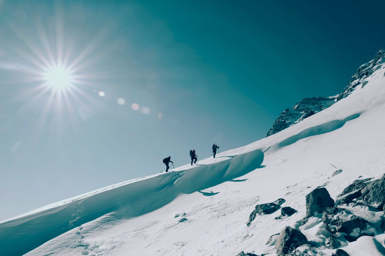 Silhouettes of hikers climbing a bright snow-covered ridge under a clear blue sky